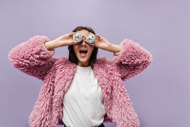 Funny stylish woman in cool t-shirt and fashionable pink fluffy sweater posing with disco balls on isolated lilac wall