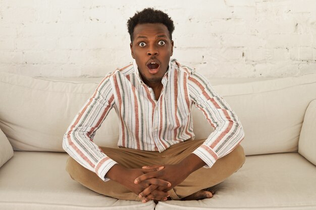 Забавно удивленный изумленный молодой африканский мужчина в повседневной одежде сидит на диване в позе полулотоса