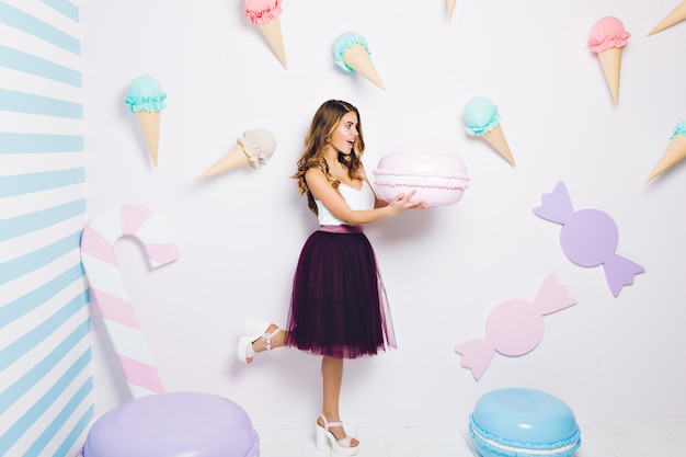 Funny surprised attractive young woman holding huge macaron among ice cream. Happy sweet moments, expressing positivity, diet concept, summer time.