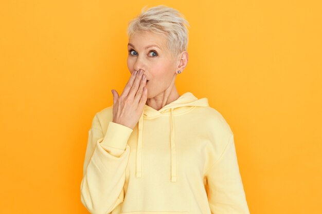 Funny surprised mature woman in hoodie covering mouth with hand, having amazed astonished facial expression, confused with intriguing amazing news. Cute retired lady expressing astonishment