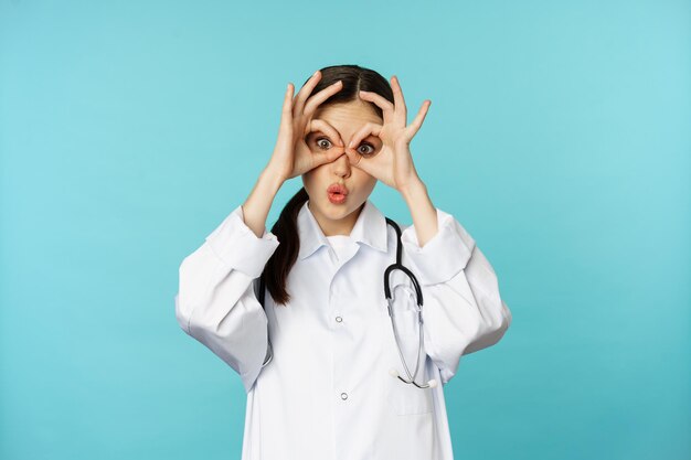 재미있는 치료사, 여의사는 괜찮은 모습을 보여주고 쌍안경은 눈에 아무런 제스처도 하지 않고 웃고, 장난을 치고 재미를 느끼며 파란 배경 위에 서 있습니다.