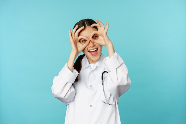 Funny therapist, girl doctor showing okay, binoculars zero gesture on eyes and smiling, fooling around and having fun, standing over blue background.