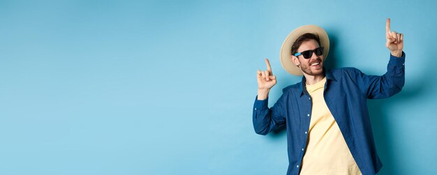 Funny tourist dancing on vacation pointing fingers up wearing summer hat and sunglasses blue backgro