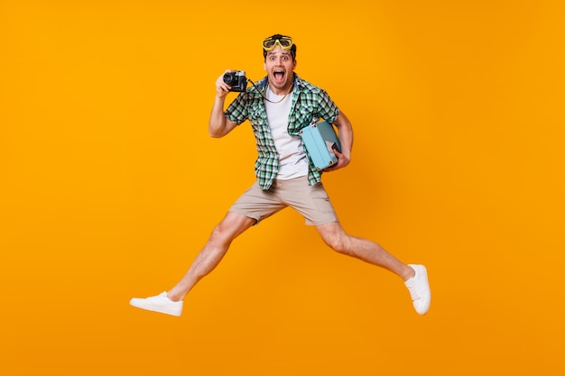 Funny tourist guy in summer outfit holding retro camera and blue suitcase. man in diving mask jumping on orange space