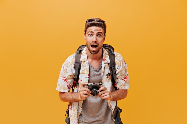 Забавный турист с голубыми глазами в солнечных очках, модной летней рубашке и серой клетчатой футболке смотрит в камеру на оранжевой стене