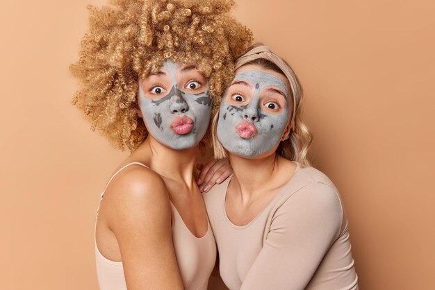 Забавные две женщины-модели держат губы округлыми, делают забавную гримасу, наносят питательные глиняные маски на лицо для омоложения и ухода за телом, одетые небрежно изолированы на коричневом фоне Концепция красоты