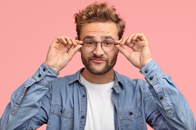 Funny unshaven man has thick beard, keeps both hands on rim of glasses, has curious look while listens something interesting