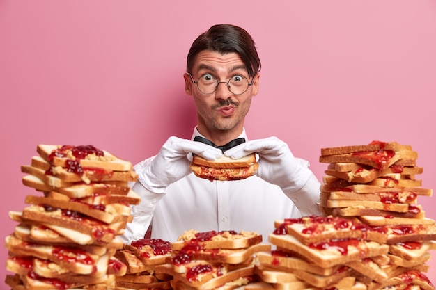 Funny waiter in snow white shirt and gloves, feels hungry after long work, holds delicious sandwich, surrounded by many toasts, wears round spectacles, doesnt keep to diet, has good appetite