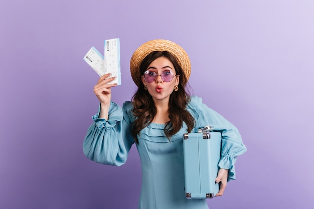 Смешная женщина в канотье и сиреневых очках смотрит в изумлении, показывая свои билеты на самолет и синий ретро чемодан.