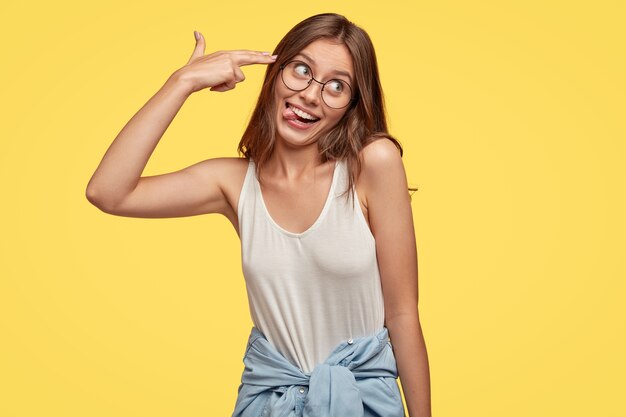 Funny woman foolishes indoor, makes finger gun gesture in temple, sticks out tongue, pretends commiting suicide, dressed in casual clothes, isolated over yellow wall, has comic expression