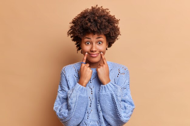 funny woman makes smile with fingers forces herself to have positive expression wears casual sweater poses against brown wall