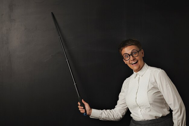 Funny woman teacher with smiling stupid face in round glasses happily shows something with her pointer on chalk board on black