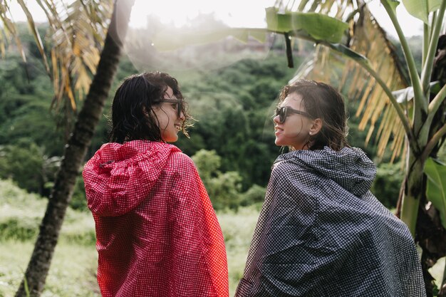 Funny women with wet hair looking at each other while walking around forest. Outdoor photo of female tourists in raincoats standing on nature.