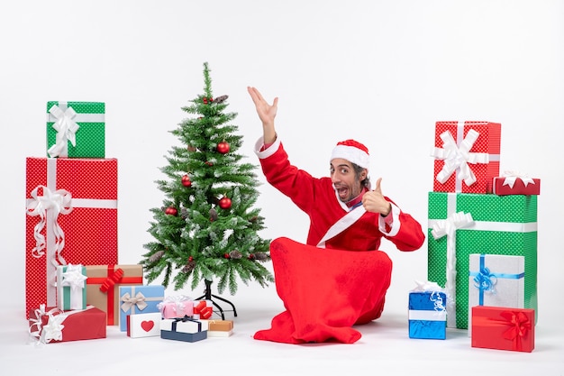 Забавный молодой человек в костюме Санта-Клауса с подарками и украшенной елкой сидит на земле, указывая вверх, делая идеальный жест на белом фоне