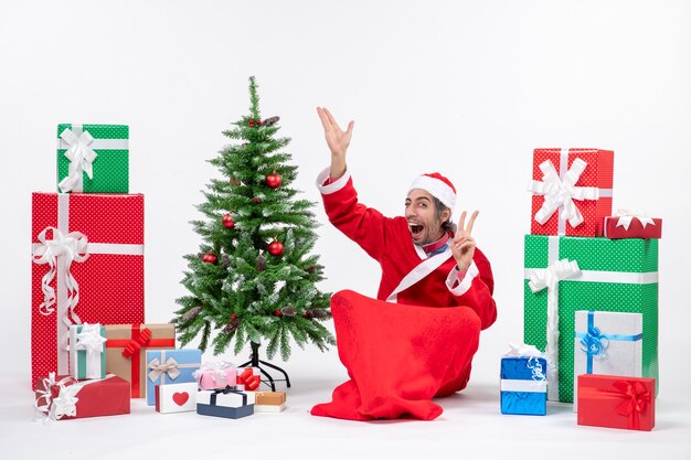 Funny young adult dressed as Santa claus with gifts and decorated Christmas tree sitting on the ground pointing above making victory gesture on white background