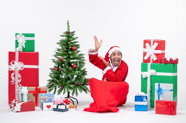 Забавный молодой человек в костюме Санта-Клауса с подарками и украшенной елкой сидит на земле, указывая вверху на белом фоне