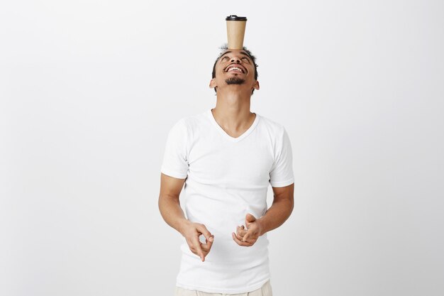 Funny young african-american guy in white t-shirt balancing coffee cup on forehead, fooling around