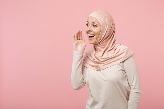 スタジオでピンクの背景に分離されたポーズをとってヒジャーブの軽い服を着た面白い若いアラビアのイスラム教徒の女性。人々の宗教的なイスラムのライフスタイルの概念。コピースペースをモックアップします。彼女の手の後ろに秘密をささやく。