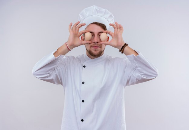 白い炊飯器の制服と白い壁に彼の目の前に有機卵を保持している帽子を身に着けている面白い若いひげを生やしたシェフの男