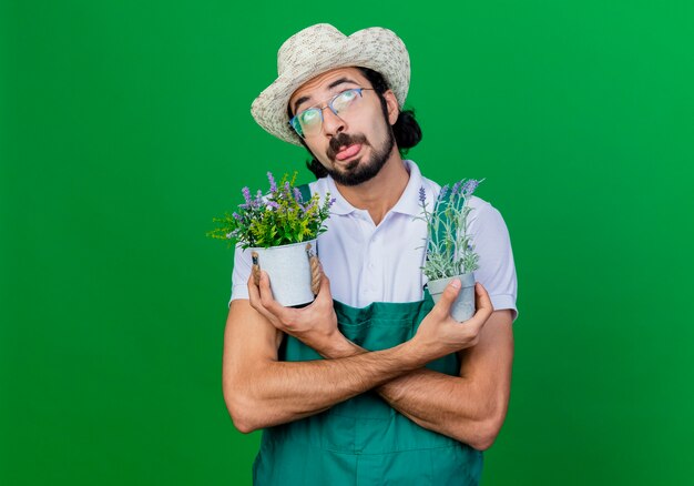 Funny young bearded gardener man wearing jumpsuit and hat holding potted plants looking up sticking out tongue 