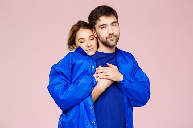 Funny young beautiful couple wearing one jacket embracing smiling over light pink wall