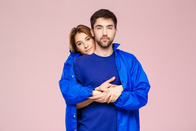 Funny young beautiful couple wearing one jacket embracing smiling over light pink wall