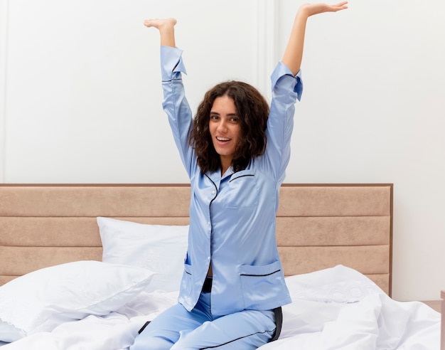 Funny young beautiful woman in blue pajamas sitting on bed happy and positive enjoying weekend in bedroom interior 