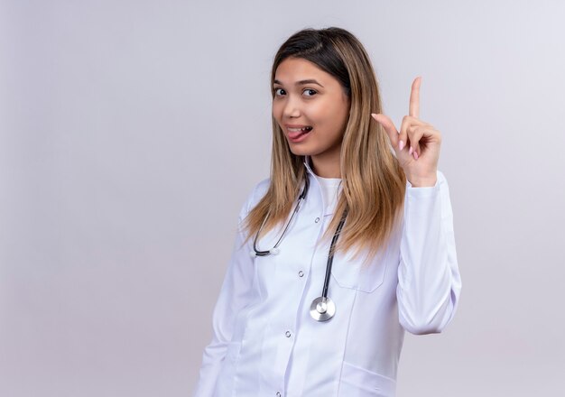 Забавная молодая красивая женщина-врач в белом халате со стетоскопом, высунувший язык, указывая указательным пальцем вверх