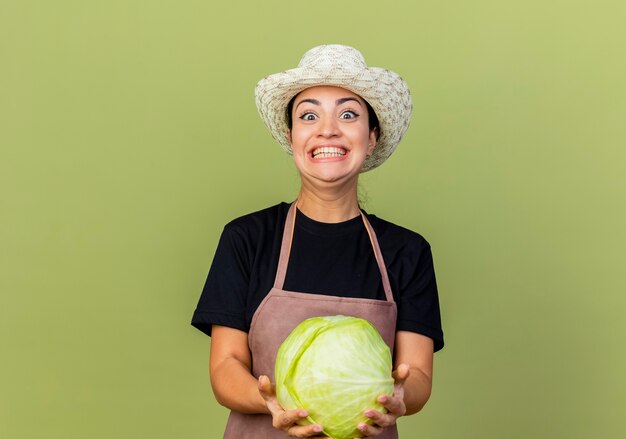 Funny young beautiful woman gardener in apron and hat holding cabbage looking at front smiling cheerfully standing over light green wall