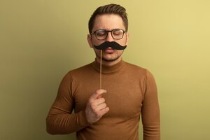 Free photo funny young blonde handsome man wearing glasses holding fake mustache on stick above lips looking down at mustache isolated on olive green wall