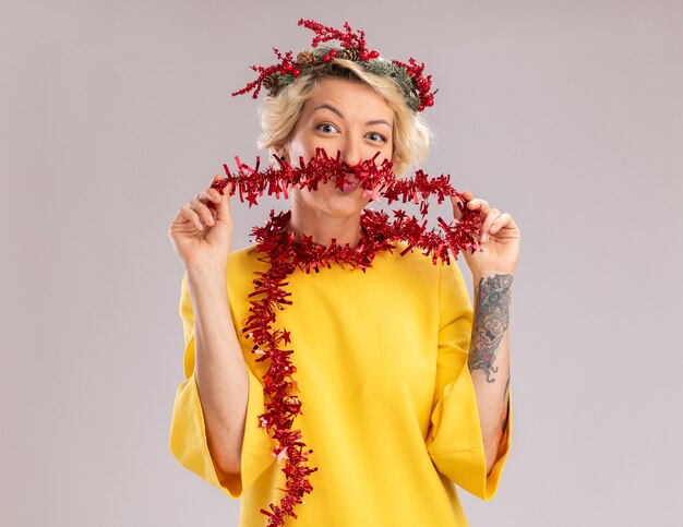 Free photo funny young blonde woman wearing christmas head wreath and tinsel garland around neck looking  making mustache with tinsel garland pursing lips isolated on white wall with copy space