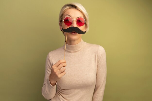 Funny young blonde woman wearing sunglasses holding fake mustache on stick above lips looking at front isolated on olive green wall with copy space