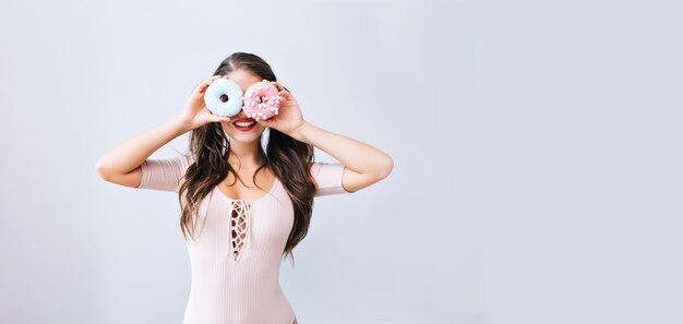 Funny young girl with long hair holding colorful donuts against her eyes. Attractive teenage having fun with sweets, delicious. Brightful lifestyle.