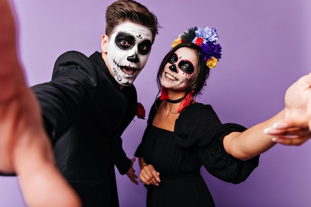 Funny young guy and his girlfriend make selfie with smiles on their faces. Portrait of naughty couple with Halloween makeup in purple studio.