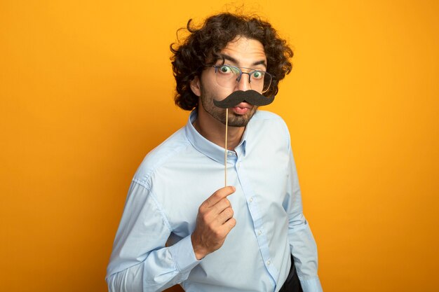 Divertente giovane uomo caucasico bello con gli occhiali mantenendo baffi finti sul bastone sopra le labbra che guarda l'obbiettivo che fa gesto di bacio isolato su priorità bassa arancione con lo spazio della copia