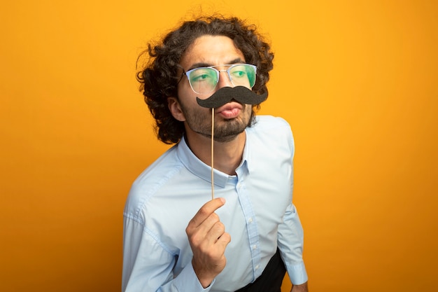 Funny young handsome man wearing glasses keeping fake mustache on stick above lips looking at side doing kiss gesture isolated on orange wall