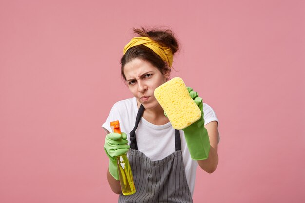 Funny young housewife wearing casual clothes, apron and protective rubber gloves obsessed with cleanliness, staring with measuring look while tidying up house until it's sparkly clean