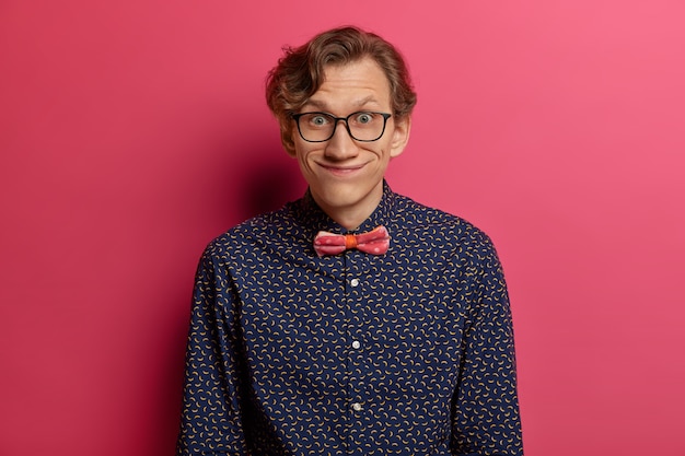 Funny young man gazes with comic expression , wears optical glasses and stylish shirt, notices something interesting, has pleasant talk with interlocutor, isolated over pink wall
