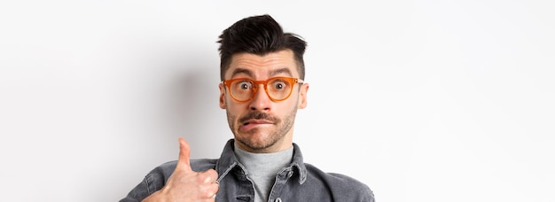 Free photo funny young man in glasses show thumb up biting lip and look indecisive standing on white background