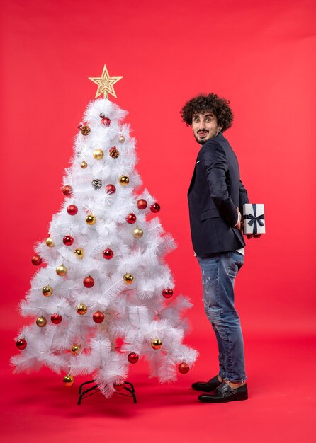 赤の右側に飾られた白いクリスマスツリーの近くに立って後ろに彼の贈り物を保持している面白い若い男