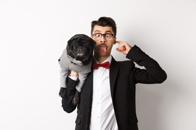 Забавный молодой человек в костюме партии, показывая язык и держа милого черного мопса на плече, празднуя с домашним животным, стоя на белом фоне.