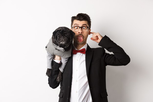 Funny young man in party suit, showing tongue and holding cute black pug on shoulder, celebrating with pet, standing over white background.