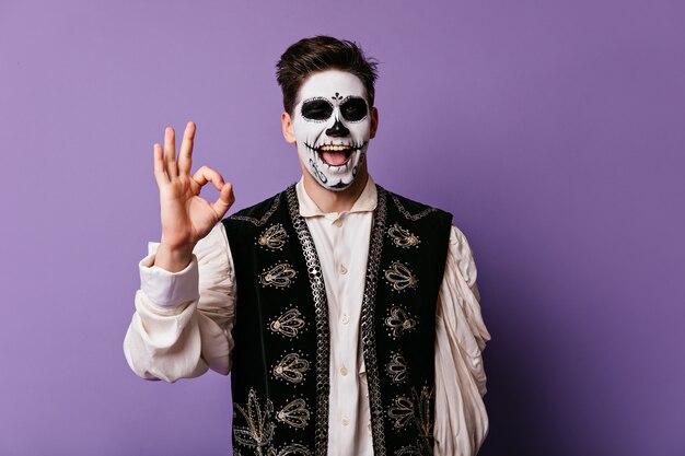 Funny young man shows sign OK. Indoor portrait of guy with face painted for Halloween.