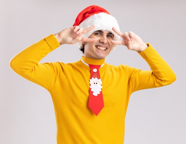 Funny young man in yellow turtleneck and santa hat with funny tie showing v-sign near eyes smiling cheerfully standing over white wall