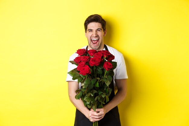 Funny young salesman in black apron holding bouquet of roses, florist laughing and standing over yellow background.