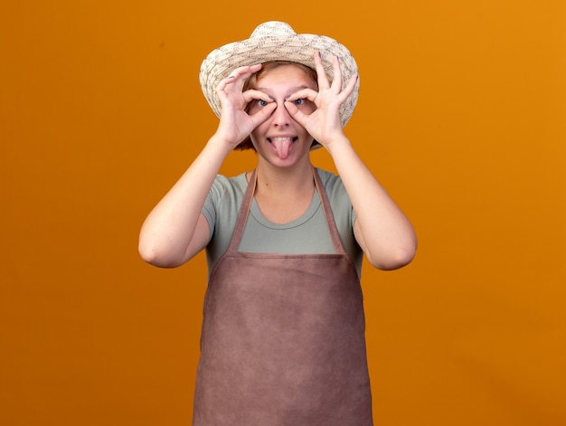 Funny young slavic female gardener wearing gardening hat stucks out tongue and looks at camera through fingers on orange