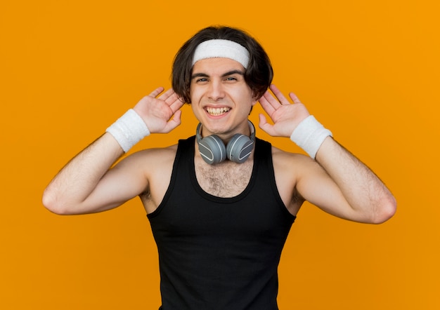 Funny young sporty man wearing sportswear and headband with headphones around neck showing ears smiling cheerfully 