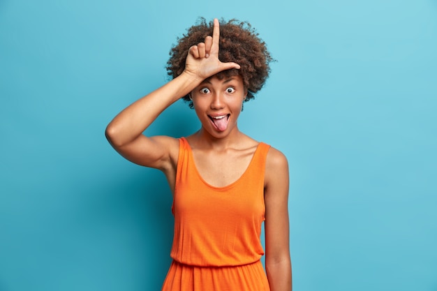 Foto gratuita gesti divertenti della giovane donna con il dito sulla fronte rendono il gesto del perdente che prende in giro la gente mostra la lingua e indossa pose di abiti arancioni contro il muro blu