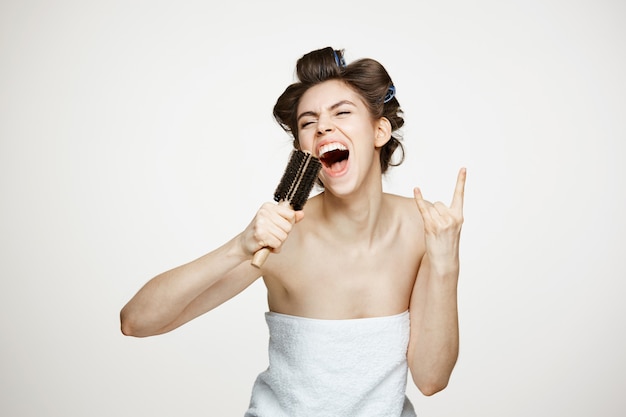 Funny young woman in hair curlers and towel rejoicing singing in comb. Beauty and spa.