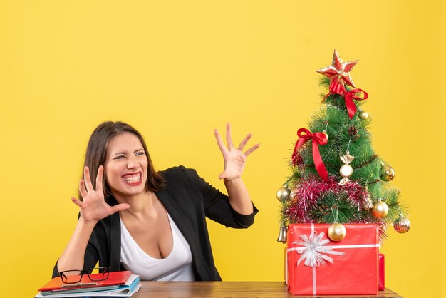 Funny young woman looking at something with surprised facial expression sitting at a table near decorated Christmas tree at office on yellow 